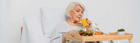 Senior patient drinking orange juice near croissant and salad on tray in hospital, bannerの写真素材