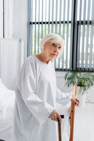 Senior woman with walking cane standing near bed in hospital wardの写真素材