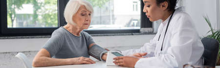 African american doctor in white coat looking at tonometer near senior patient in hospital, bannerの写真素材