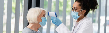 African american doctor in medical mask holding pyrometer near senior patient, bannerの写真素材