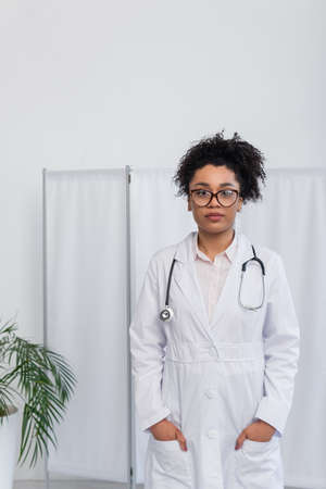 African american doctor in eyeglasses holding hands in pockets of white coatの写真素材