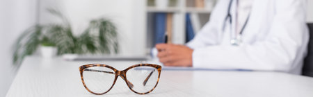 Cropped view of eyeglasses on table near blurred doctor in hospital, bannerの写真素材