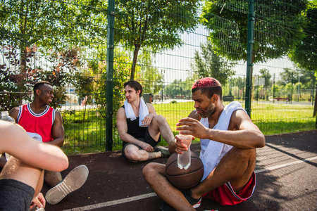 African american sportsman holding bottle of water near basketball ball and friends