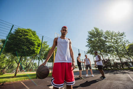 Low angle view of african american man with basketball ball looking at camera near friends on playgroundの写真素材