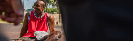 Positive african american sportsman with towel sitting near water on playground, bannerの写真素材