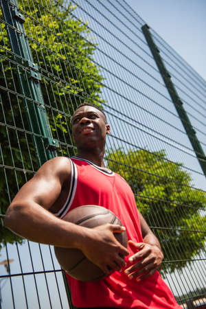 Low angle view of smiling african american sportsman holding basketball ball near fenceの写真素材