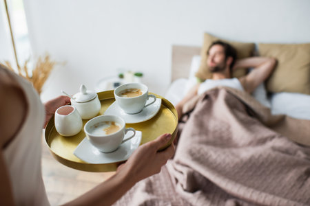 woman holding tray with cups of coffee near blurred and sleeping manの写真素材