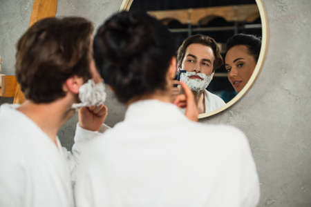 reflection in mirror of woman looking at man shaving in bathroomの写真素材