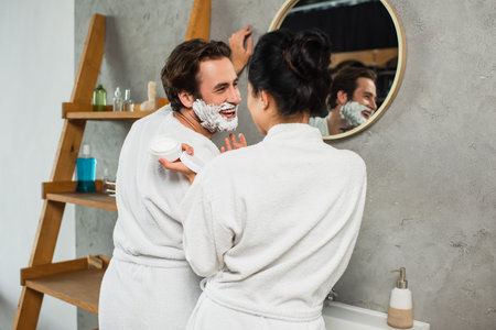 woman looking at happy man with shaving foam on faceの写真素材