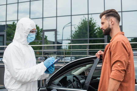 Driver standing near car and medical worker in hazmat suit writing on clipboard