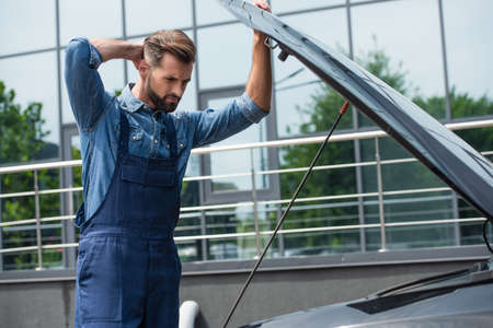 Thoughtful mechanic looking at car with open hood outdoorsの写真素材