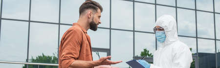 Man talking to medical worker in hazmat suit writing on clipboard outdoors, banner