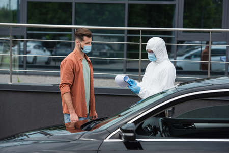 Medical worker in hazmat suit writing on clipboard near man in mask and auto outdoors