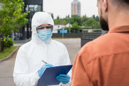 Medical worker in hazmat suit writing on clipboard near blurred man outdoorsの写真素材