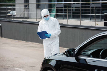 Medical worker in hazmat suit and goggles writing on clipboard near blurred car