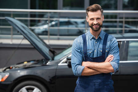 Positive mechanic in overalls looking at camera near blurred carの写真素材
