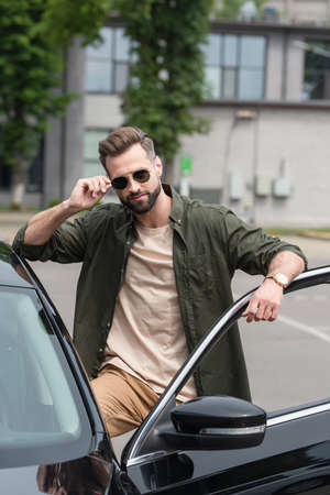 Bearded man adjusting sunglasses near auto outdoors