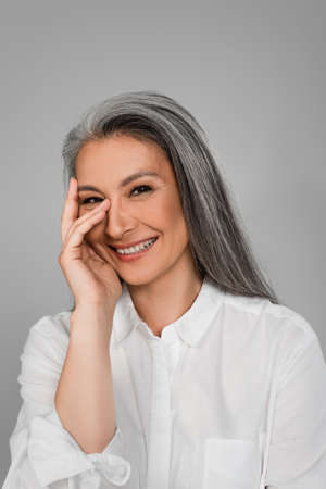 cheerful middle aged woman holding hand near face while smiling at camera isolated on grayの写真素材