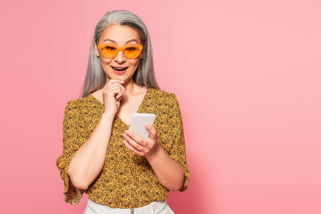 cheerful middle aged woman touching chin while using smartphone isolated on pink