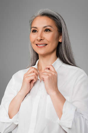 mature woman smiling while touching collar of white shirt isolated on grayの写真素材