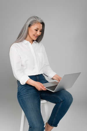 happy mature woman in white shirt typing on laptop while sitting on stool isolated on grayの写真素材