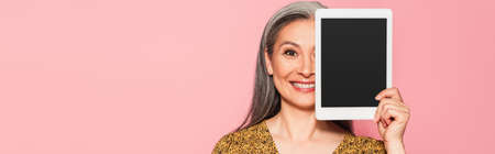 happy mature woman obscuring face with digital tablet isolated on pink, bannerの写真素材