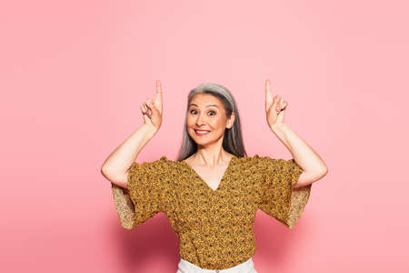 cheerful middle aged woman pointing up with fingers on pink background