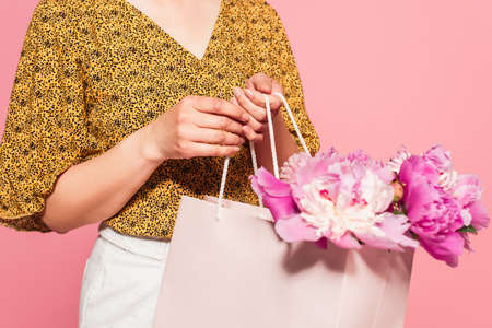 partial view of woman in stylish blouse holding shopping bag with peonies isolated on pinkの写真素材