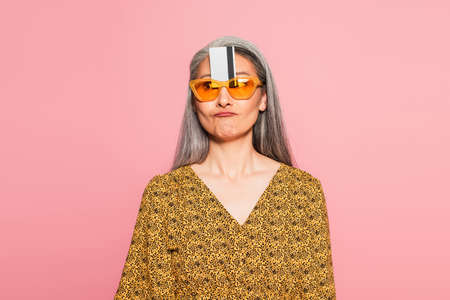 mature woman with credit card between sunglasses and forehead grimacing isolated on pink