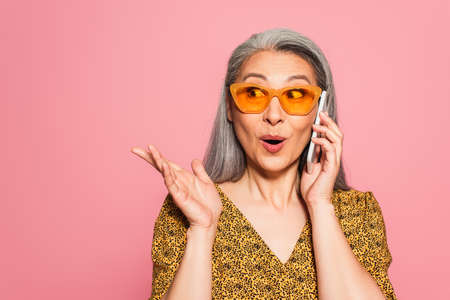 surprised middle aged woman in yellow sunglasses talking on cellphone and pointing with hand...