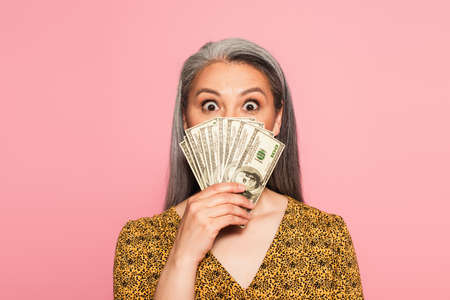 amazed middle aged woman covering face with dollar banknotes isolated on pink