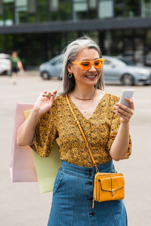cheerful woman in patterned blouse and sunglasses using cellphone while standing with shopping bags