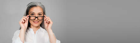 positive, gray haired woman smiling at camera while taking off eyeglasses isolated on gray, banner