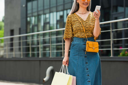 partial view of middle aged woman with shopping bags using mobile phone in city