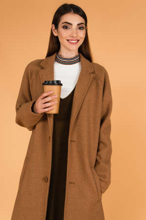 happy woman with coffee to go standing with hand in pocket of stylish coat isolated on beige