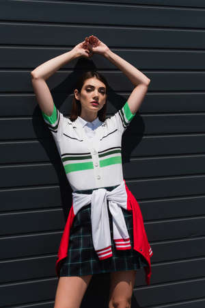 young woman in striped polo t-shirt posing near gray wall with raised hands