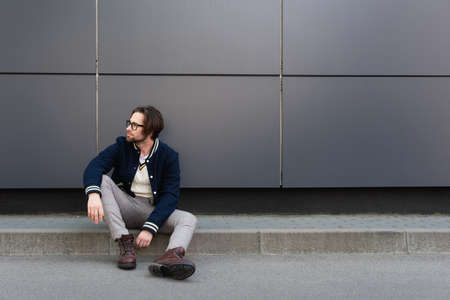 young man in trendy autumn clothes looking away while sitting on border near gray wall