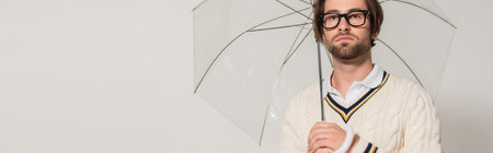 young man in white sweater and eyeglasses standing under transparent umbrella isolated on gray, bannerの写真素材