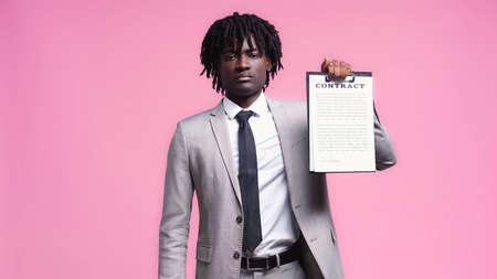 african american man in suit holding clipboard with contract lettering isolated on pink
