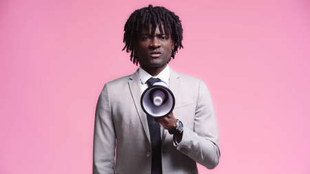 Angry african american businessman speaking into loudspeaker isolated on pinkの写真素材