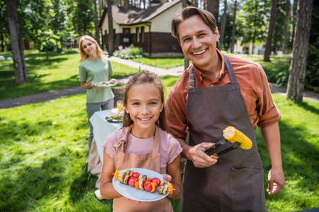Smiling father and kid in aprons holding grilled vegetables outdoorsの写真素材