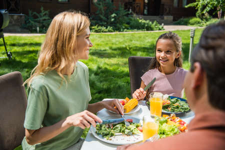 Smiling kid looking at mother with cutlery near food and orange juice outdoorsの写真素材