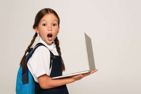 amazed schoolgirl with open mouth looking at camera while holding laptop isolated on grayの写真素材