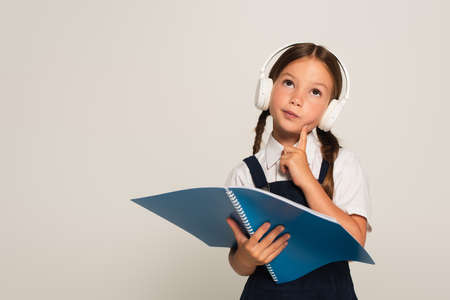 thoughtful schoolkid in headphones looking up while holding notebook isolated on grayの写真素材