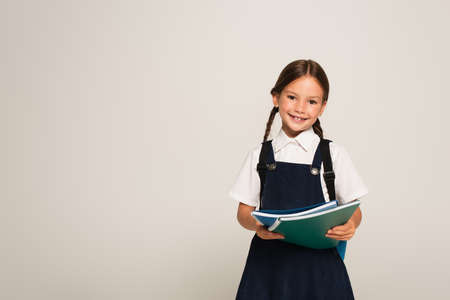 positive schoolchild smiling at camera while holding notebooks isolated on grayの写真素材