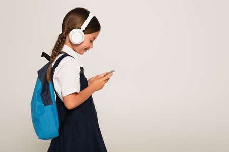 side view of cheerful schoolkid in headphones using smartphone isolated on grayの写真素材
