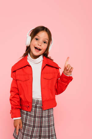 cheerful girl in orange jacket and headphones pointing with finger isolated on pinkの写真素材
