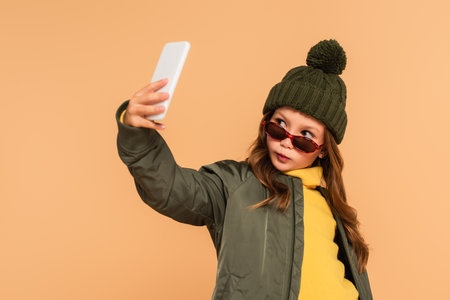 stylish kid in autumn clothes taking selfie on smartphone isolated on beigeの写真素材