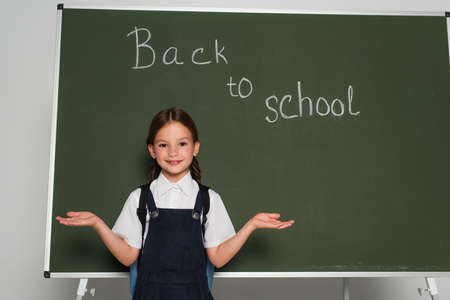 cheerful schoolgirl pointing with hands near chalkboard with back to school lettering on gray