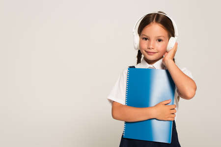 smiling schoolkid adjusting headphones while holding notebook isolated on grayの写真素材